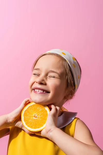 Lächelndes Mädchen mit geschlossenen Augen, das orange halb isoliert auf rosa hält — Stockfoto