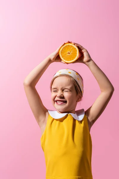 Smiling girl with closed eyes holding orange half above head isolated on pink — Stock Photo