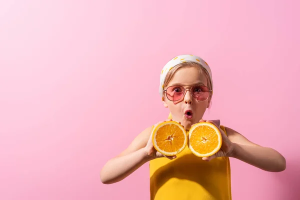 Shocked girl in headscarf and sunglasses holding orange halves on pink — Stock Photo