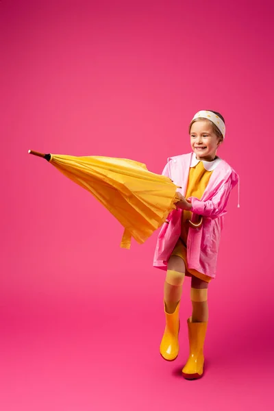 In voller Länge glückliches Mädchen in Regenmantel und Gummistiefeln mit Regenschirm auf purpurrotem Grund — Stockfoto