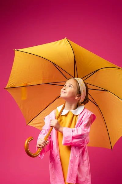 Zufriedenes Mädchen im Regenmantel steht unter gelbem Regenschirm isoliert auf purpurrotem Grund — Stockfoto