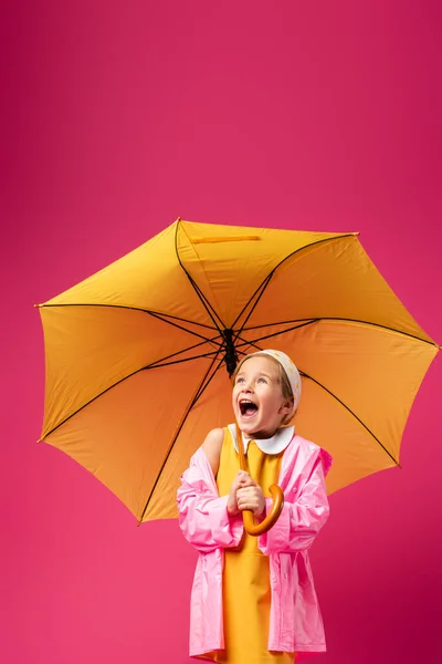 Erstauntes Mädchen im Regenmantel steht unter Regenschirm auf purpurrotem Grund — Stockfoto
