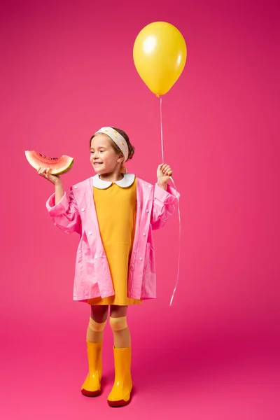 Volle Länge des glücklichen Mädchens im Regenmantel mit Luftballon und aufgeschnittener Wassermelone auf Karminrot — Stockfoto
