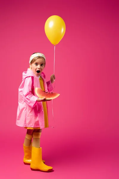 Volle Länge des erstaunten Mädchens im Regenmantel mit Luftballon und Wassermelone in Scheiben auf Karminrot — Stockfoto