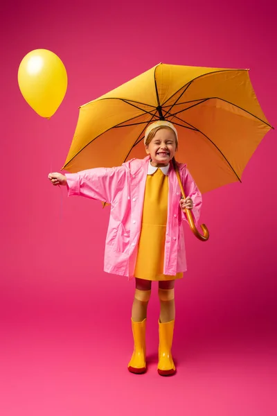 In voller Länge aufgeregtes Mädchen im Regenmantel mit Luftballon und gelbem Regenschirm auf Purpur — Stockfoto
