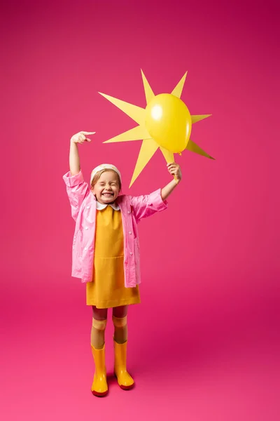 In voller Länge aufgeregtes Mädchen in Regenmantel und Gummistiefeln mit dekorativer Sonne und Luftballon auf Purpur — Stockfoto