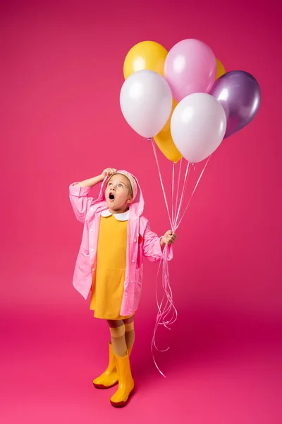 Longitud completa de chica sorprendida en impermeable y botas de goma sosteniendo globos en carmesí - foto de stock