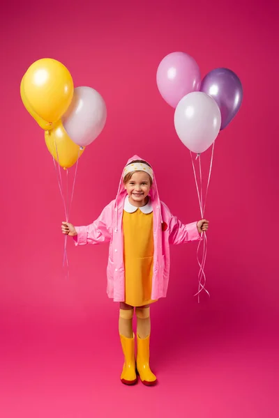 In voller Länge glückliches Mädchen in Regenmantel und Gummistiefeln mit Luftballons auf purpurrotem Grund — Stockfoto
