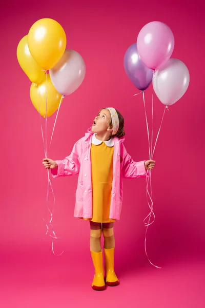 In voller Länge staunende Mädchen in Regenmantel und Gummistiefeln, die Luftballons auf purpurrotem Grund halten — Stockfoto