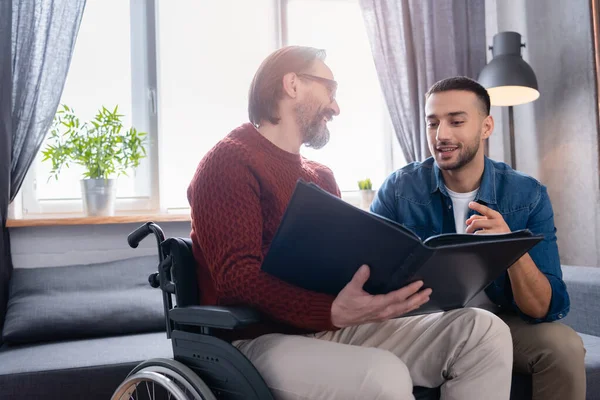 Happy hispanic man pointing with finger at photo album in hands of smiling disabled father — Stock Photo