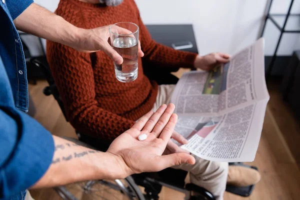 Vista recortada del hombre dando agua y píldora a padre discapacitado sentado en silla de ruedas con periódico, enfoque selectivo - foto de stock