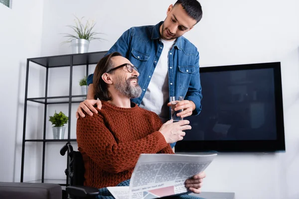 Joven hispano dando agua a feliz padre discapacitado sentado en silla de ruedas con periódico - foto de stock