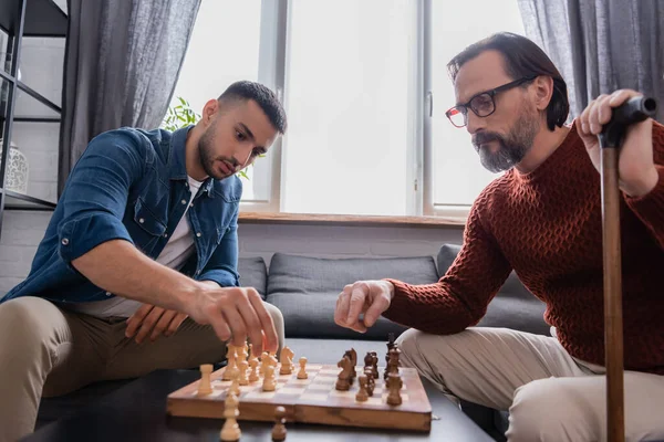 Serio uomo barbuto con bastone da passeggio giocare a scacchi con figlio ispanico a casa — Foto stock