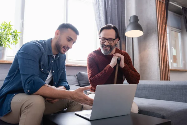 Young hispanic man pointing at laptop near cheerful father sitting with walking stick — Stock Photo