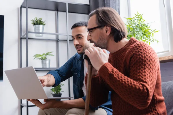 Young hispanic man pointing at laptop near father with walking stick — Stock Photo
