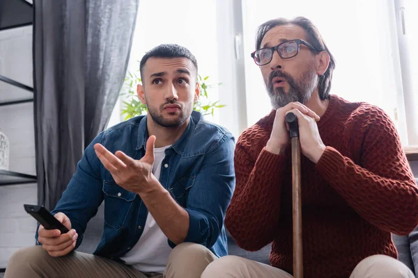 Young hispanic man pointing with hand while watching tv near amazed father — Stock Photo