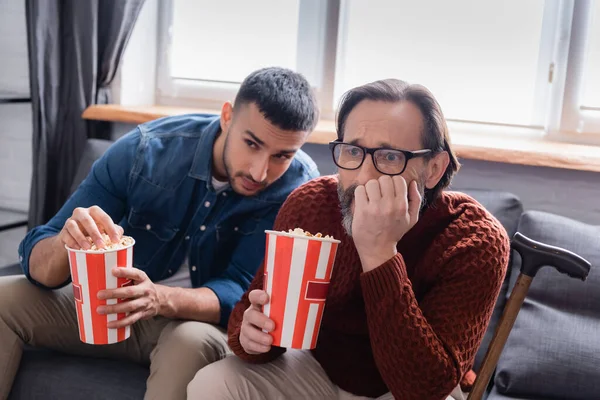 Preocupado hombre sosteniendo palomitas de maíz mientras ve la televisión con su hijo hispano en casa - foto de stock