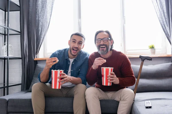 Emocionado padre interracial y el hijo riendo mientras ve la comedia y comer palomitas de maíz en casa - foto de stock