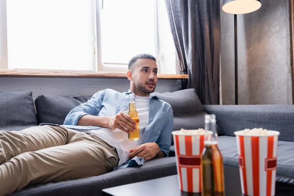 Joven hispano bebiendo cerveza y viendo la televisión mientras está acostado en el sofá - foto de stock