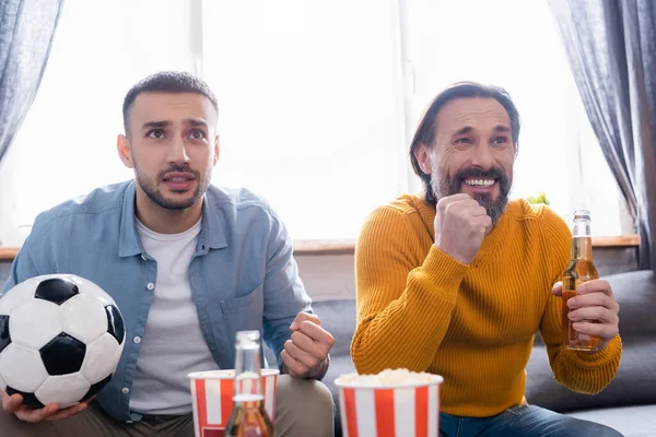 Worried interracial father and son watching football match with beer and popcorn at home — Stock Photo