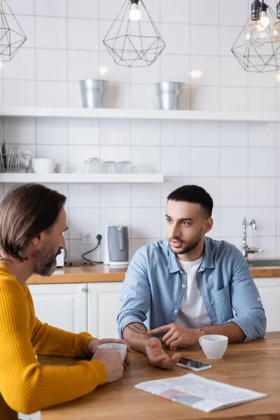 Joven hispano hijo apuntando a tatuaje en la mano durante conversación con padre en cocina - foto de stock