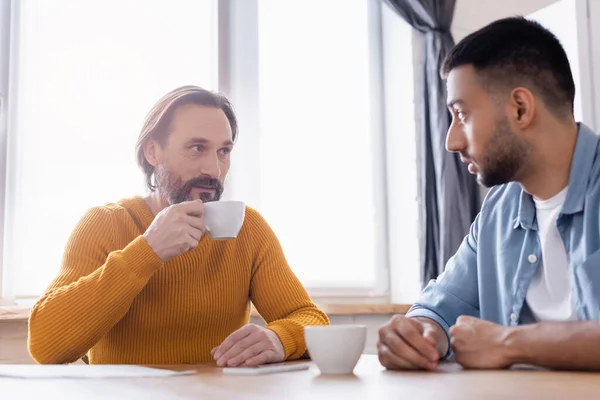 Homme barbu tenant tasse de café pendant la conversation avec son fils hispanique dans la cuisine — Photo de stock