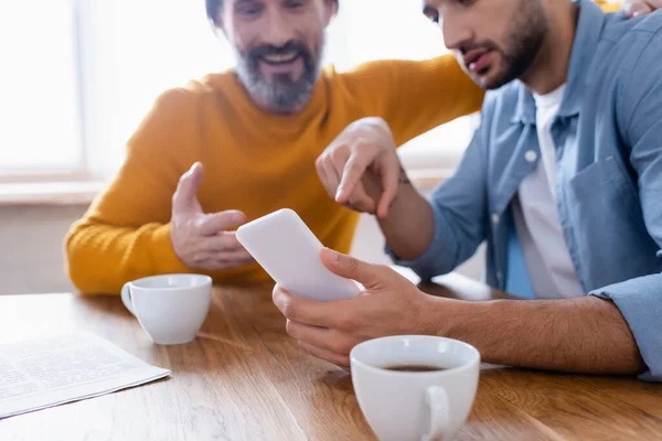 Teilbild eines hispanischen Mannes, der in der Küche neben lächelndem Vater auf sein Smartphone zeigt, verschwommener Hintergrund — Stockfoto