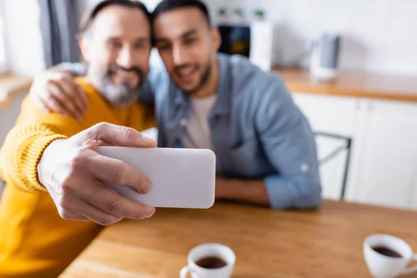 Enfoque selectivo de teléfono inteligente en la mano del hombre feliz tomando selfie con el hijo hispano sobre fondo borroso - foto de stock