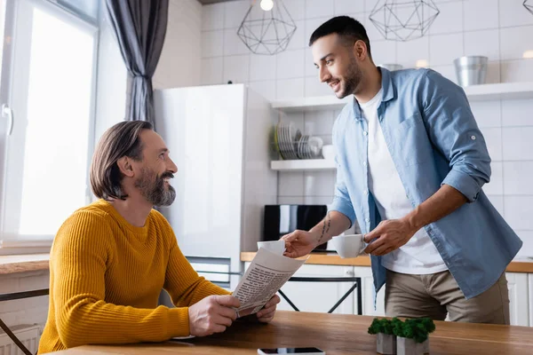 Sonriente hombre hispano sosteniendo tazas de café cerca de padre sentado con periódico en la cocina - foto de stock