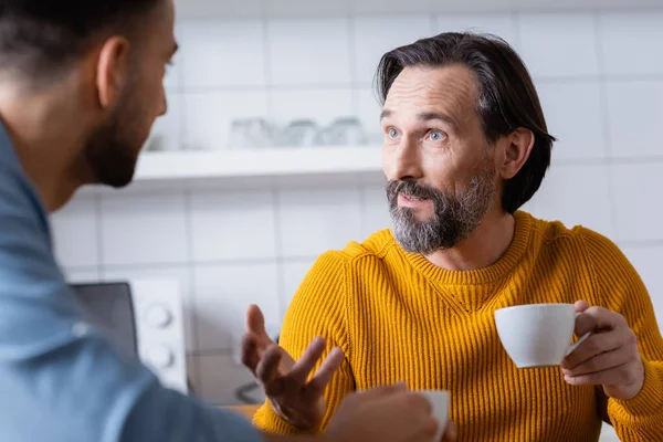 Barbudo hombre sosteniendo la taza de café y el gesto mientras habla con el hijo en la cocina, borrosa primer plano - foto de stock