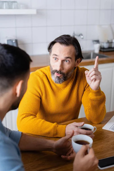 Amazed man pointing with finger while talking to son in kitchen, blurred foreground — Stock Photo