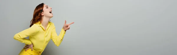 Mujer emocionada riendo y señalando con el dedo mientras está de pie con la mano en la cadera en gris, bandera - foto de stock