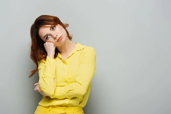 Mujer triste mirando hacia otro lado mientras está de pie con la mano cerca de la cara en gris - foto de stock