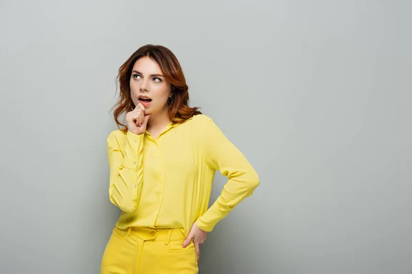 Thoughtful woman touching chin while standing with hand on hip on grey — Stock Photo