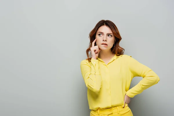 Nachdenkliche Frau beißt Lippe im Stehen mit der Hand auf der Hüfte auf grau — Stockfoto