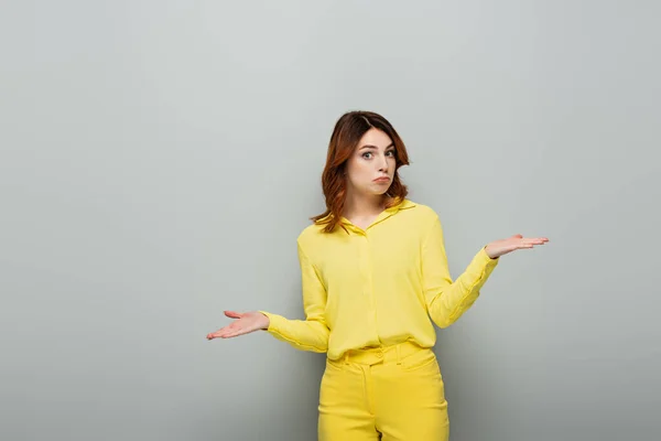 Mujer desanimada en ropa amarilla mostrando gesto de encogimiento y mirando a la cámara en gris - foto de stock