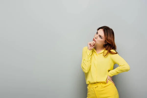 Задумчивая женщина в желтой одежде, стоящая с рукой на бедре на сером — стоковое фото