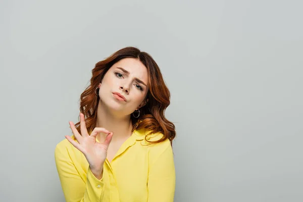 Skeptical woman looking at camera while showing ok gesture on grey — Stock Photo