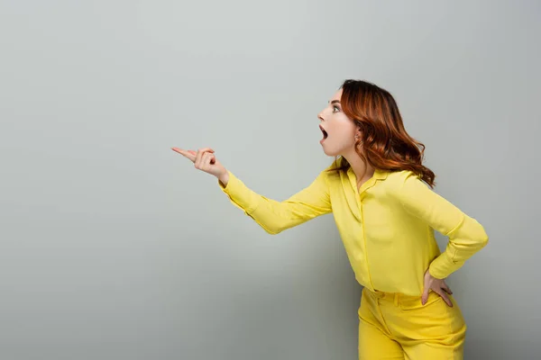 Schockierte Frau zeigt mit dem Finger, während sie mit der Hand auf der Hüfte auf grau steht — Stockfoto