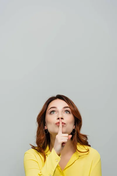 Mujer positiva mirando hacia arriba mientras muestra gesto shh aislado en gris - foto de stock