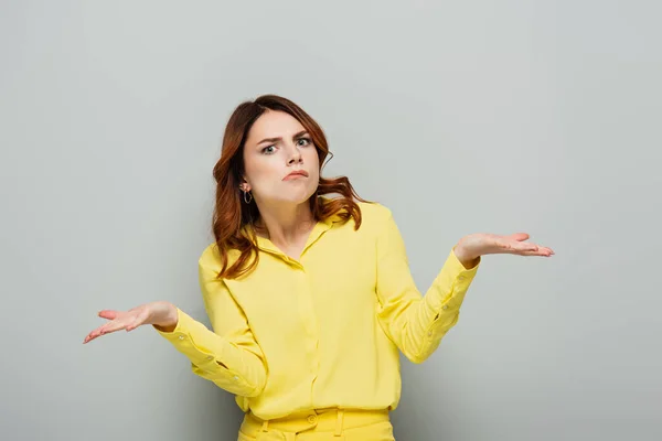 Confused woman looking at camera while showing shrug gesture on grey — Stock Photo