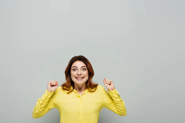 Mujer alegre sonriendo a la cámara mientras está de pie con los puños cerrados en gris - foto de stock