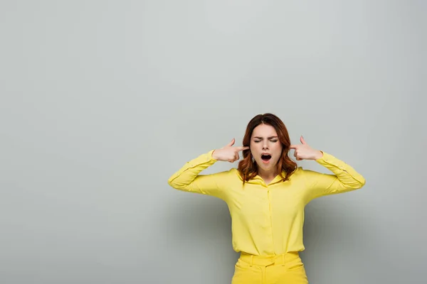 Irritated woman plugging ears while standing with closed eyes on grey — Stock Photo