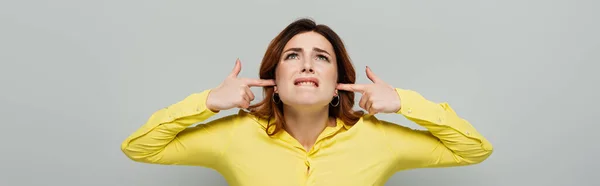 Mujer disgustada tapando las orejas mientras mira hacia arriba en gris, bandera - foto de stock