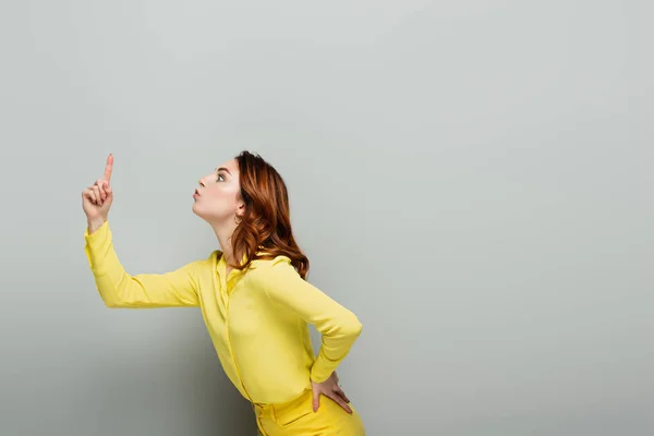 Mujer emocionada apuntando hacia arriba con el dedo mientras está de pie con la mano en la cadera en gris - foto de stock