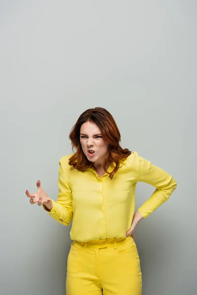 Mulher irritada com grimace irritado mostrando gesto assustador enquanto está de pé com a mão no quadril no cinza — Fotografia de Stock