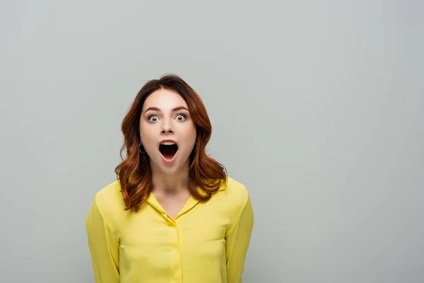 Femme étonnée en chemise jaune regardant la caméra tout en se tenant debout avec la bouche ouverte isolé sur gris — Photo de stock