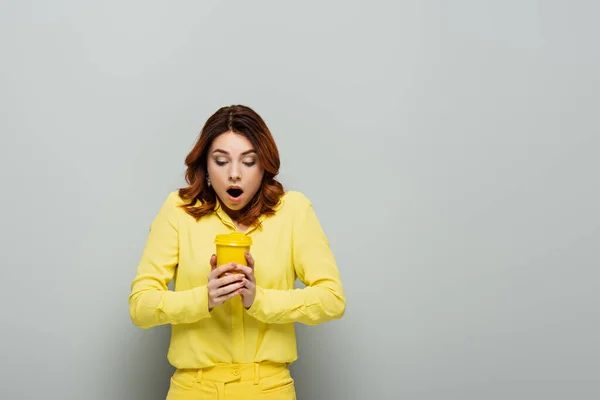 Femme choquée en chemisier jaune tenant café chaud en tasse en papier jaune sur gris — Photo de stock