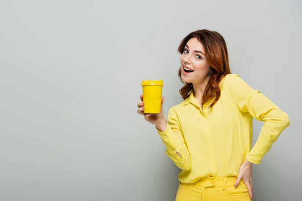 Cheerful woman holding yellow paper cup while standing with hand on hip on grey — Stock Photo