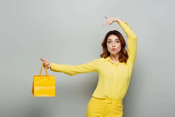 Excited woman pointing at yellow shopping bag while looking at camera on grey — Stock Photo